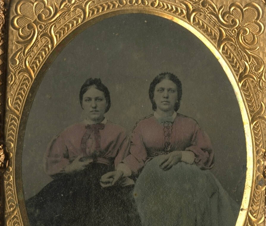 Black and white ambrotype photographic portrait of two seated women. Some hand coloring visible on their clothing including bright pink shirts.
